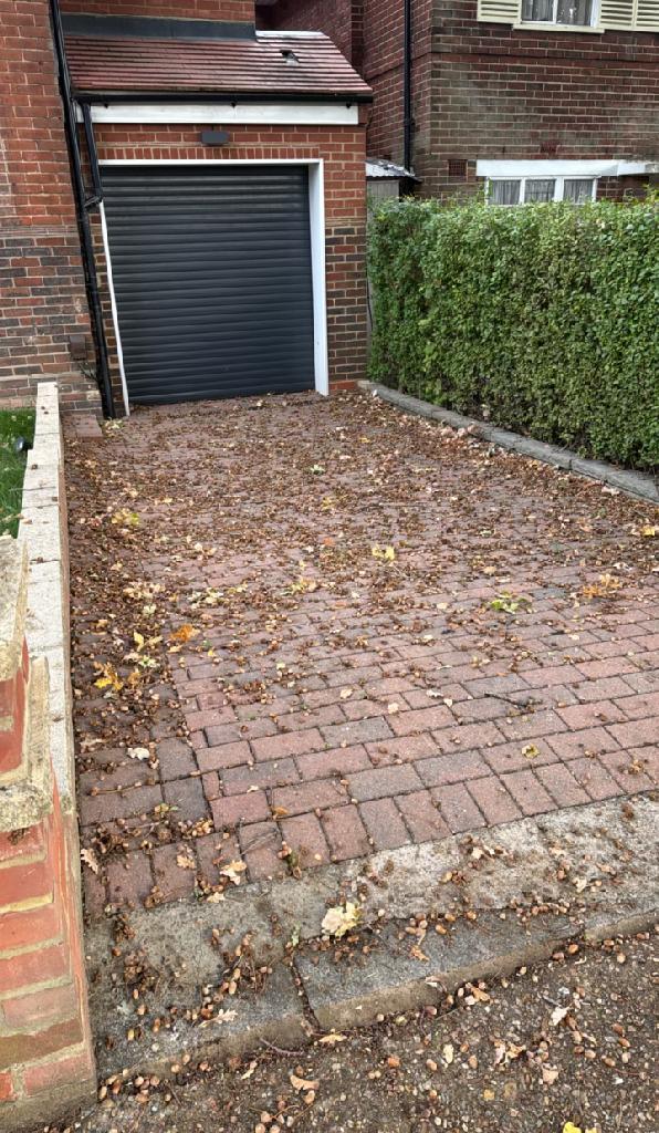 Paved front area beside a garage with leaf litter and weed growth across paving blocks, with bare grey concrete borders.